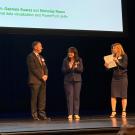 Three people stand on a stage in front of a projection screen during a presentation